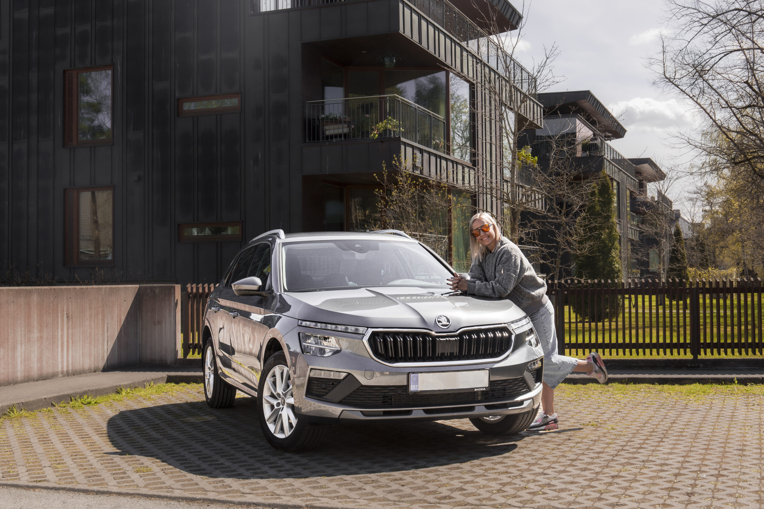 Pilkas Skoda kamiq iš mobire veiklos nuomos asortimento prie kurio stovis į kėbulą pasirėmusi moteris